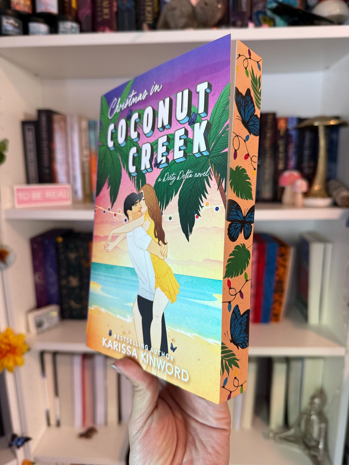 A book titled 'Christmas in Coconut Creek' and 'A Dirty Delta Novel' by Karissa Kinwood displayed in front of a bookshelf
