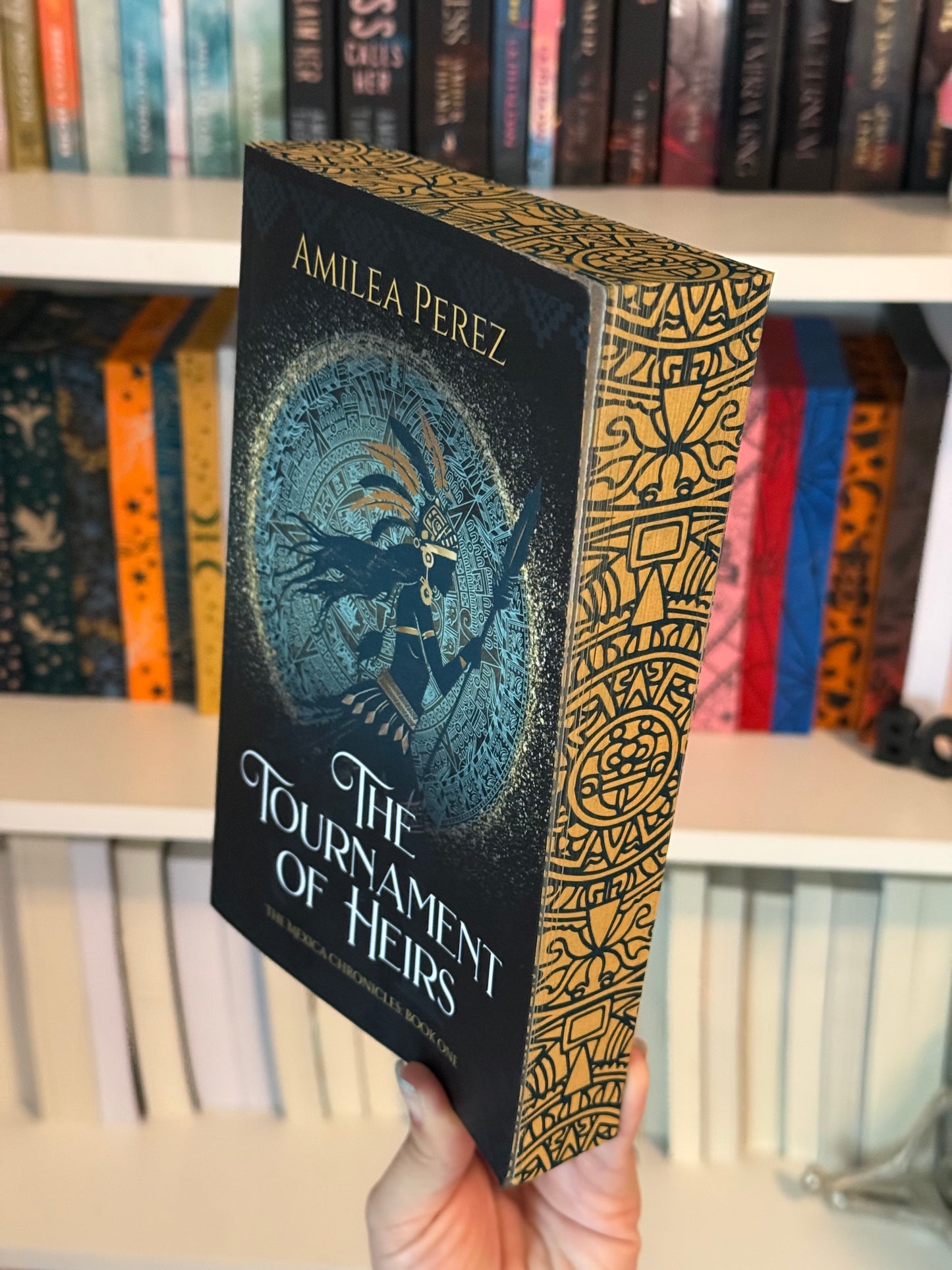 A book titled 'The Tournament of Heirs and The Mexica Chronicles: Book One' by Amilea Perez displayed in front of a bookshelf