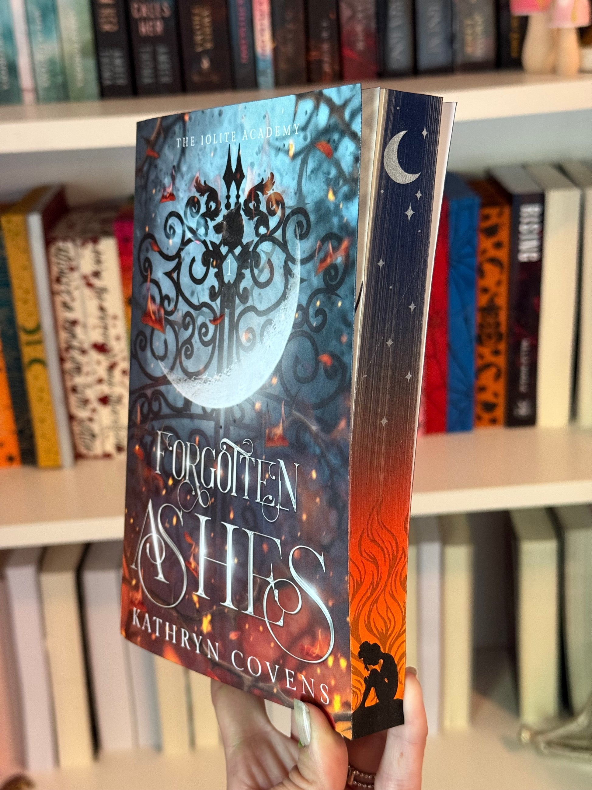 A book titled 'Forgotten Ashes' by Kathryn Covens displayed in front of a bookshelf
