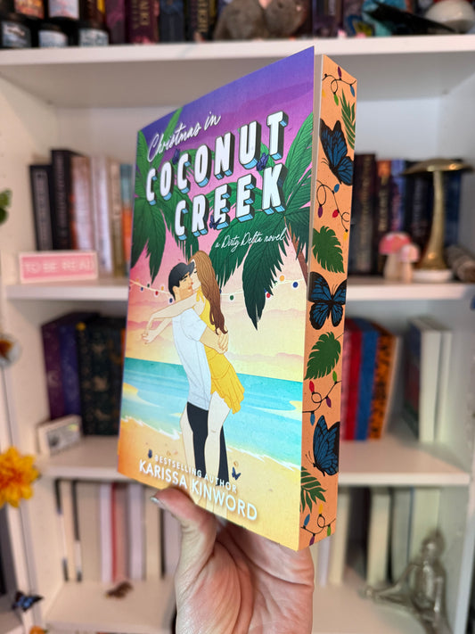 A book titled 'Christmas in Coconut Creek' and 'A Dirty Delta Novel' by Karissa Kinwood displayed in front of a bookshelf