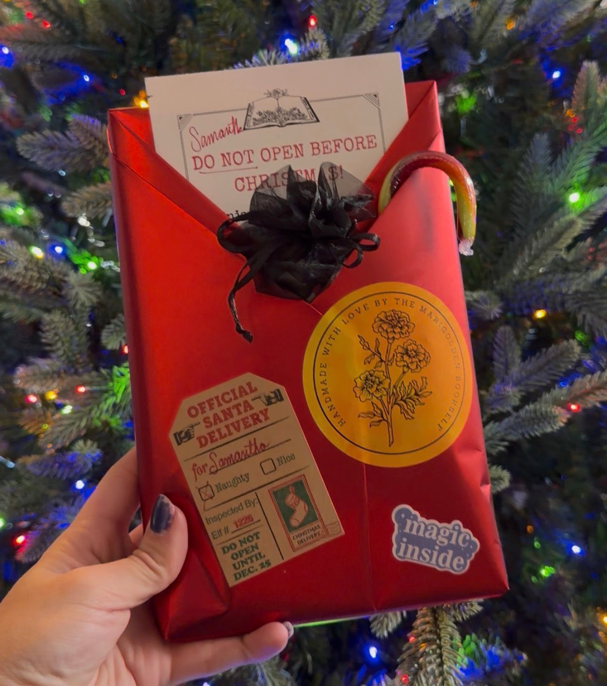 Red gift bag with festive text and a black bow, held in front of a decorated Christmas tree.