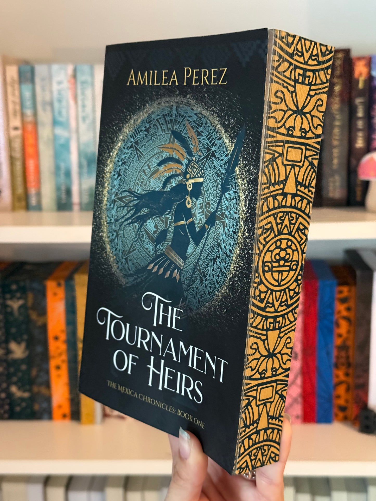 A book titled 'The Tournament of Heirs and The Mexica Chronicles: Book One' by Amilea Perez displayed in front of a bookshelf