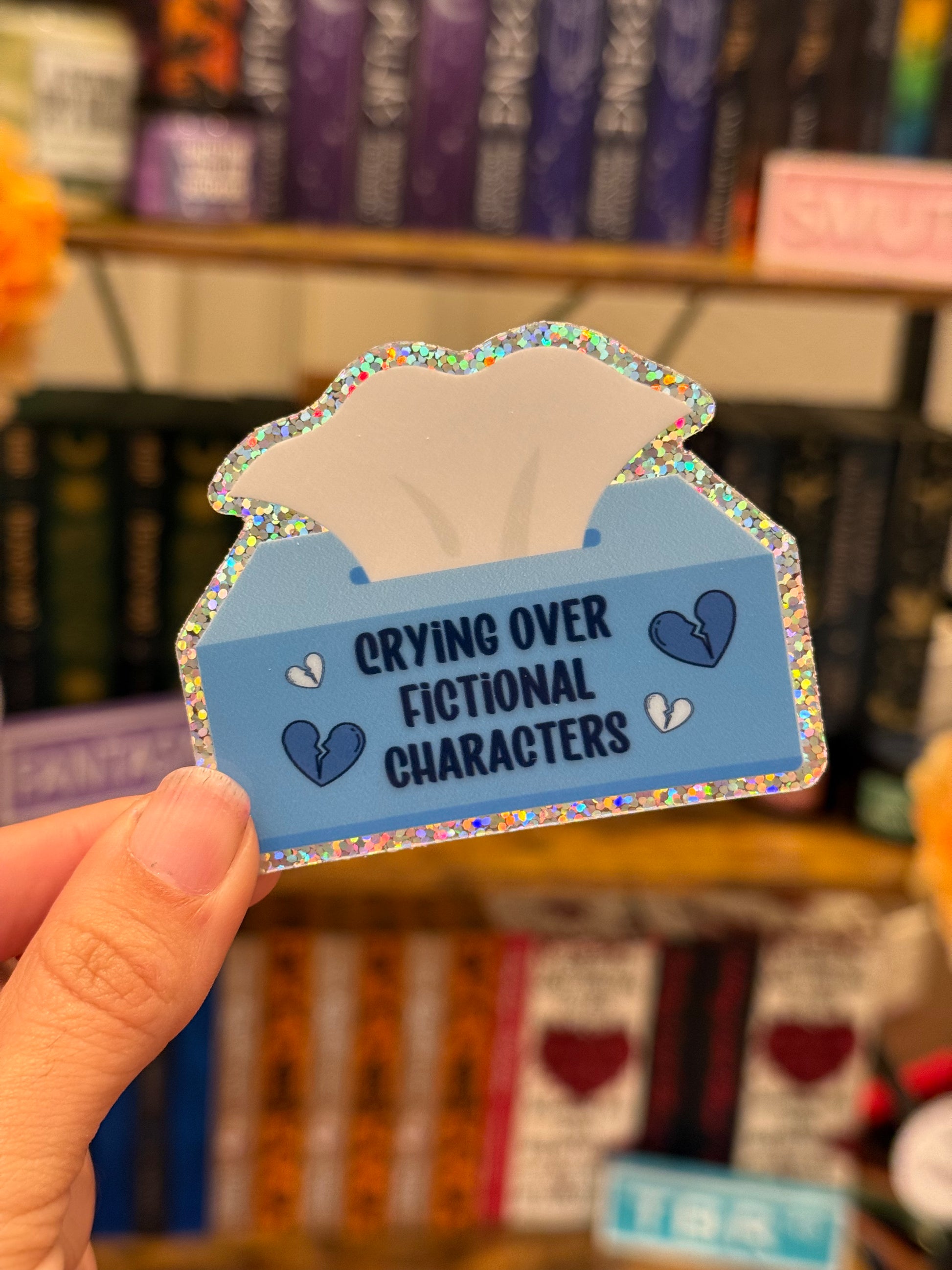 Sticker shaped like a tissue box with text 'Crying over fictional characters' held in front of a bookshelf.