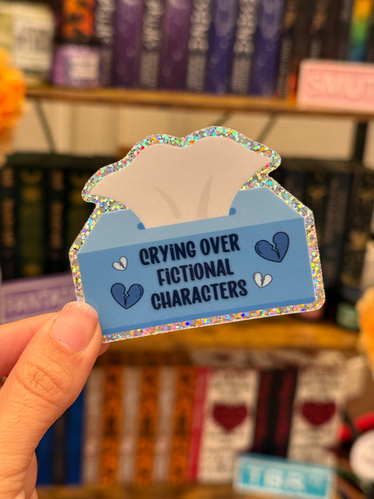 Sticker shaped like a tissue box with text 'Crying over fictional characters' held in front of a bookshelf.